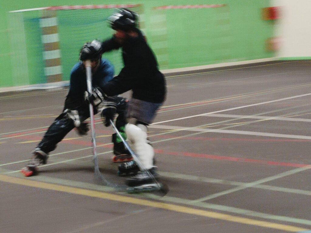 roller hockey ,2 joueurs en action avec crosses , casques et protections de joueur Le premier debout ,tire le second en position de freinage jambe droite en avant . Dans une salle de handball avec un revêtement au sol en goudron